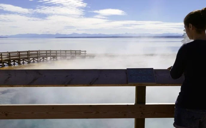 A young woman looks over a steaming pool and a lake from the railes of a boardwalk